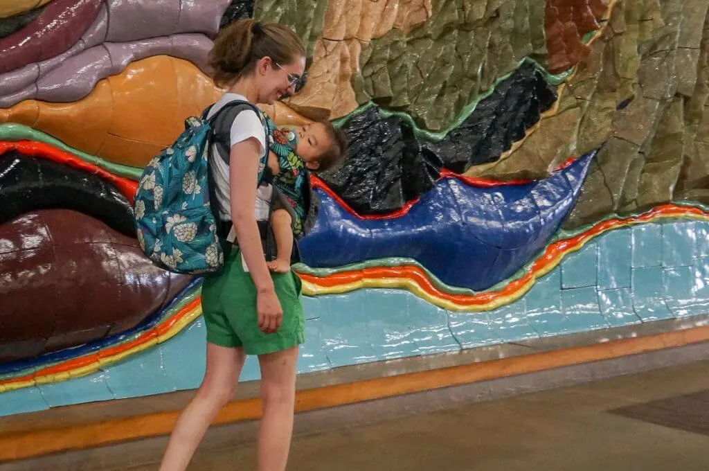 Image of a mom wearing a Hawaiian print backpack while wearing a baby in a baby carrier at the Lihue Airport on Kauai.