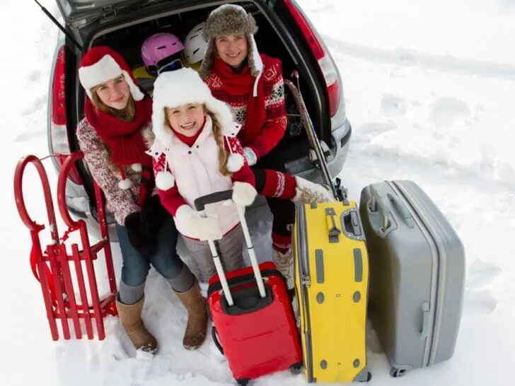 Find out the top winter travel packing tips by top family travel blog Marcie in Mommyland. Image of a family wearing holiday sweaters with luggage in the snow.