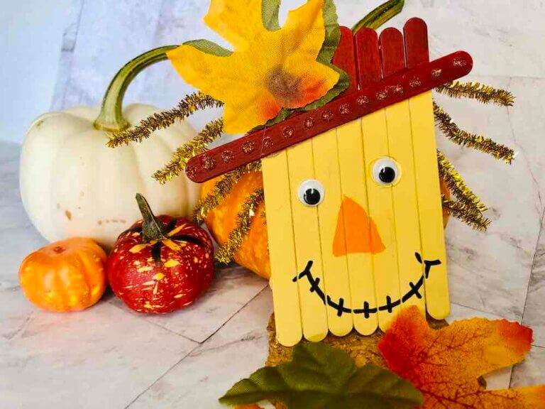 Learn how to make a Popsicle Stick Scarecrow Craft from top Seattle blog Marcie in Mommyland. Image of a popsicle stick scarecrow face in front of mini pumpkins