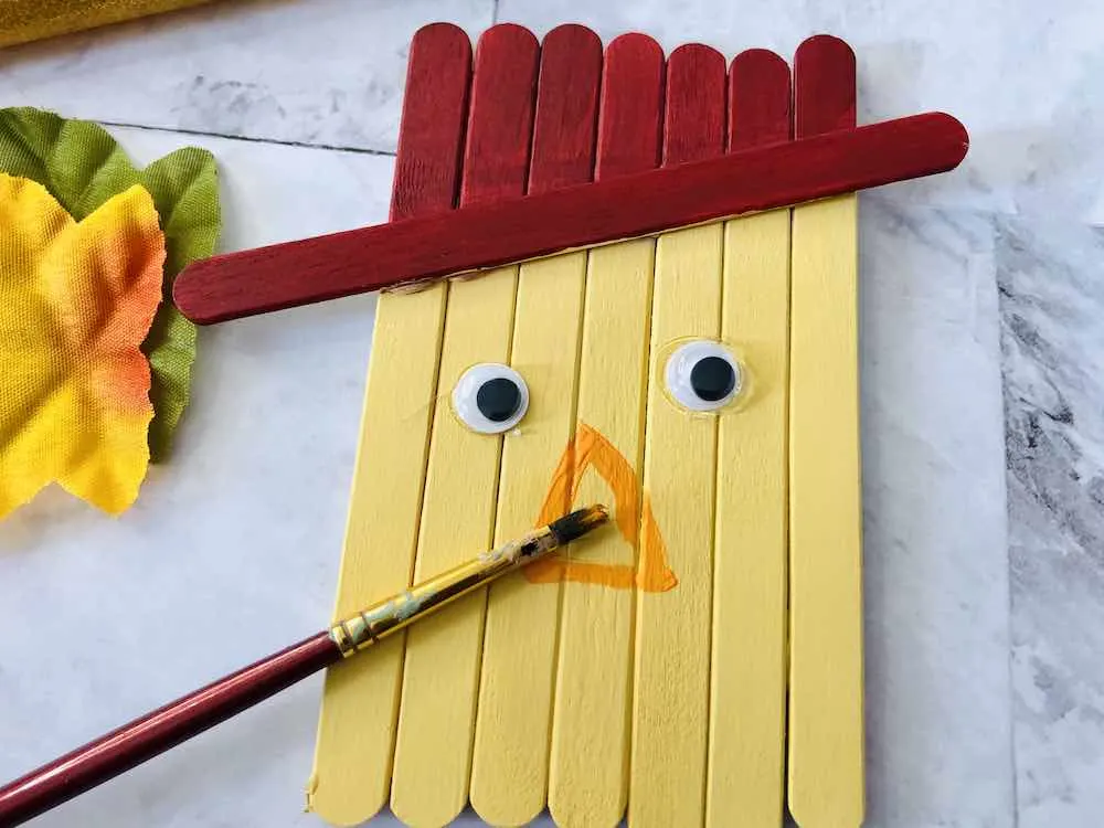 How to make a popsicle stick scarecrow craft step 6. Image of someone painting an orange nose on the scarecrow