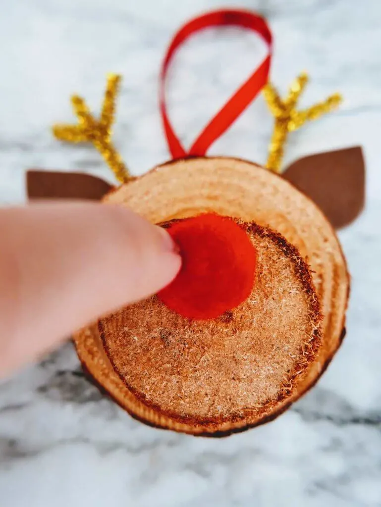 How to make a DIY reindeer ornament step 12. Image of someone gluing a red pom pom onto the reindeer ornament.