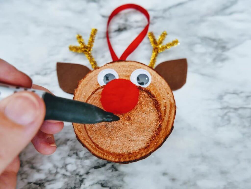 How to make a DIY reindeer ornament step 14. Image of someone drawing a mouth on their wood reindeer ornament.