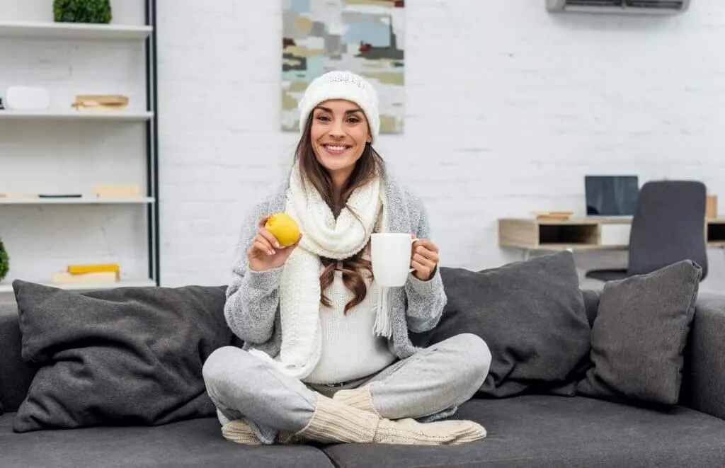 Throw on some extra layers to stay warm at home this winter. Image of a woman at home wearing a hat, scarf, sweater, and socks