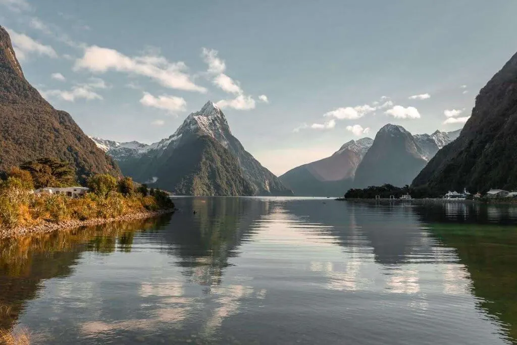 Milford Sound is a South Island New Zealand must see attraction. Image of still water with mountains surrounding and a partly cloudy sky.