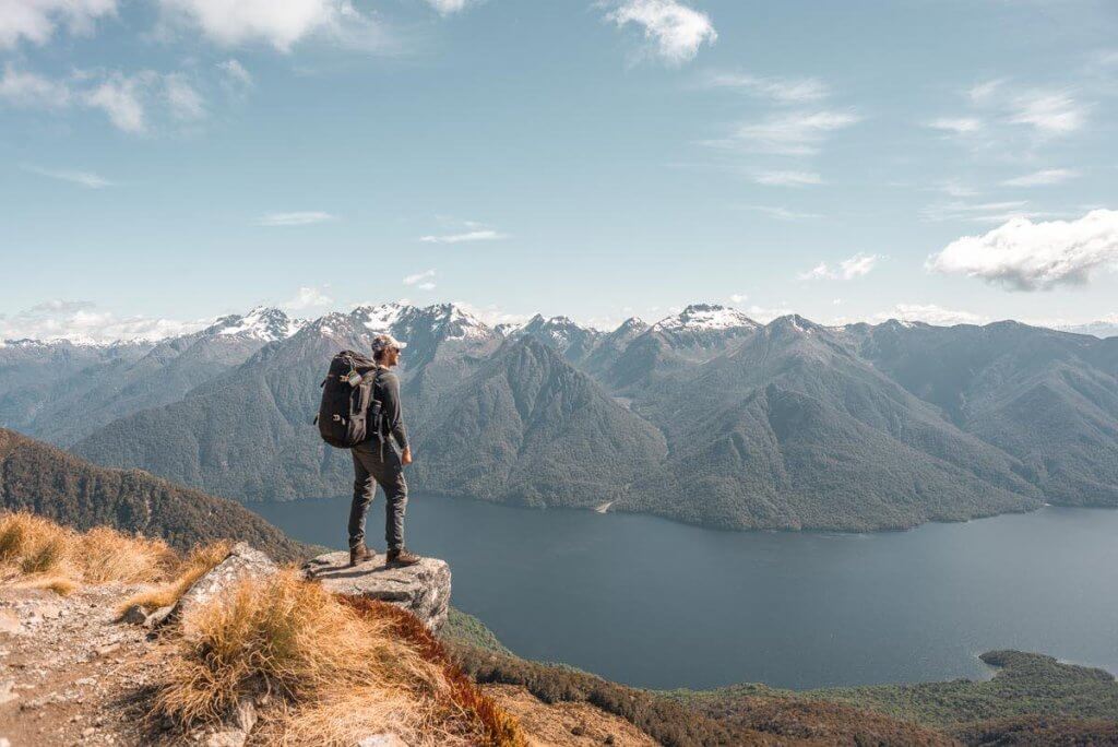 Kepler Track in Te Anau is one of the top places to visit in South Island NZ. Image of a man with a hiking backpack standing at a lookout with water and mountains in the background.