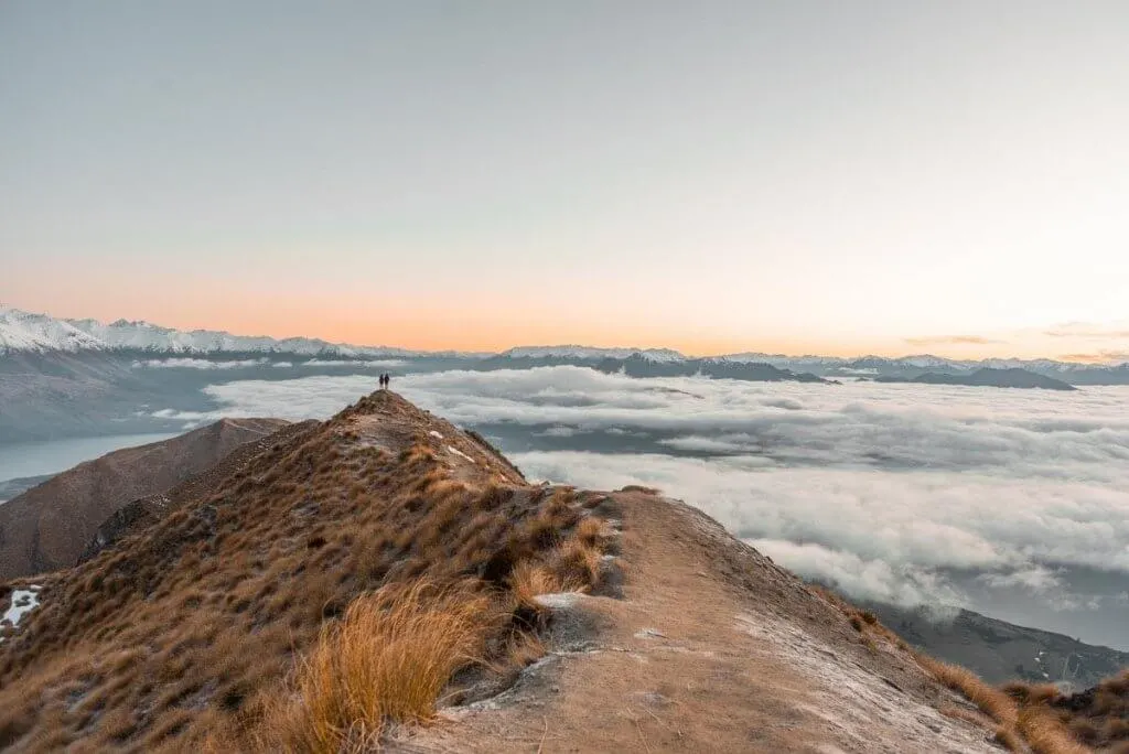 Roy's Peak in Wanaka is one of the best places to visit in South Island New Zealand. Image of a golden mountain peak at sunrise with a pinkish sky and low clouds.