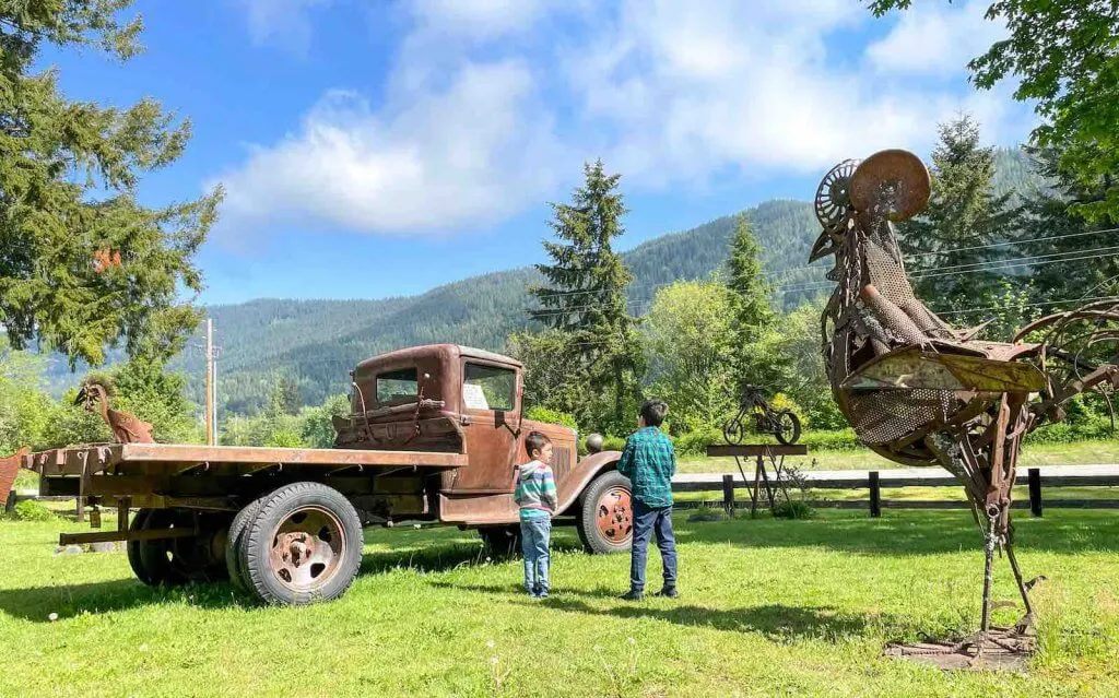 The Recycled Spirits of Iron Sculpture Park is a fun Mt Rainier attraction for families. Image of two boys standing near an old truck and giant chicken statue.