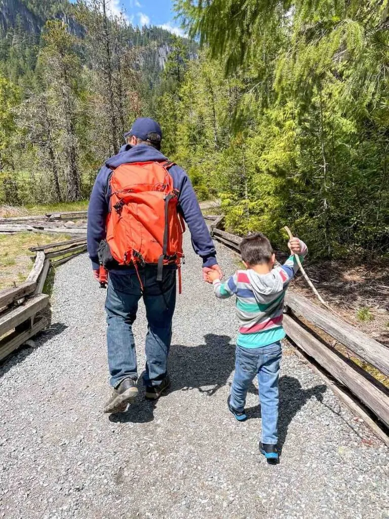 The Trail of Shadows is one of the best Mt Rainier hikes for kids. Image of a dad and son walking on a gravel path at Mount Rainier National Park.