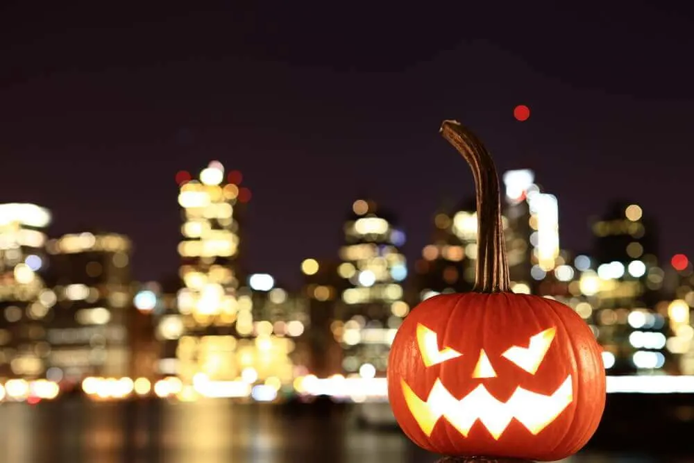 New York City is one of the best places to visit for Halloween. Image of a jack-o-lantern with nighttime lights of NYC in the background.