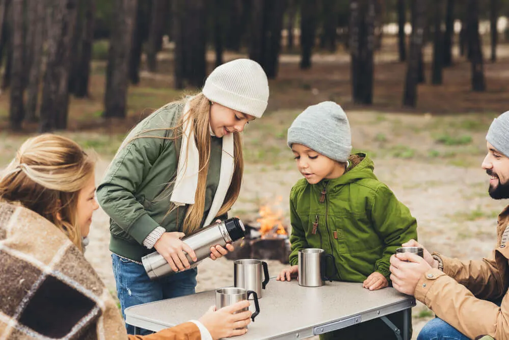 Make sure to check out this fall camping packing list for families so you don't forget anything. Image of a family pouring hot cocoa into thermoses while on a fall camping trip.
