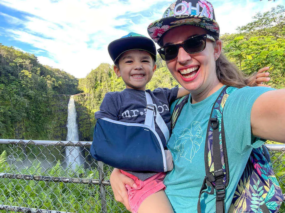 Image of a mom and a boy with his arm in a sling taking a selfie in front of a waterfall on the Big Island of Hawaii.