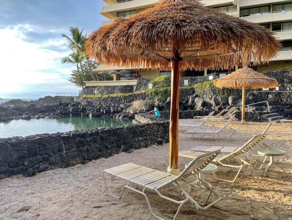 Image of white chaise lounges under thatched umbrellas on the beach in Kona Hawaii.