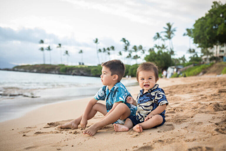 Find out the top 25 things to do on Maui with kids by a mom who's visited more than 10 times! Image of two boys wearing Aloha shirts sitting on a beach in Maui Hawaii.