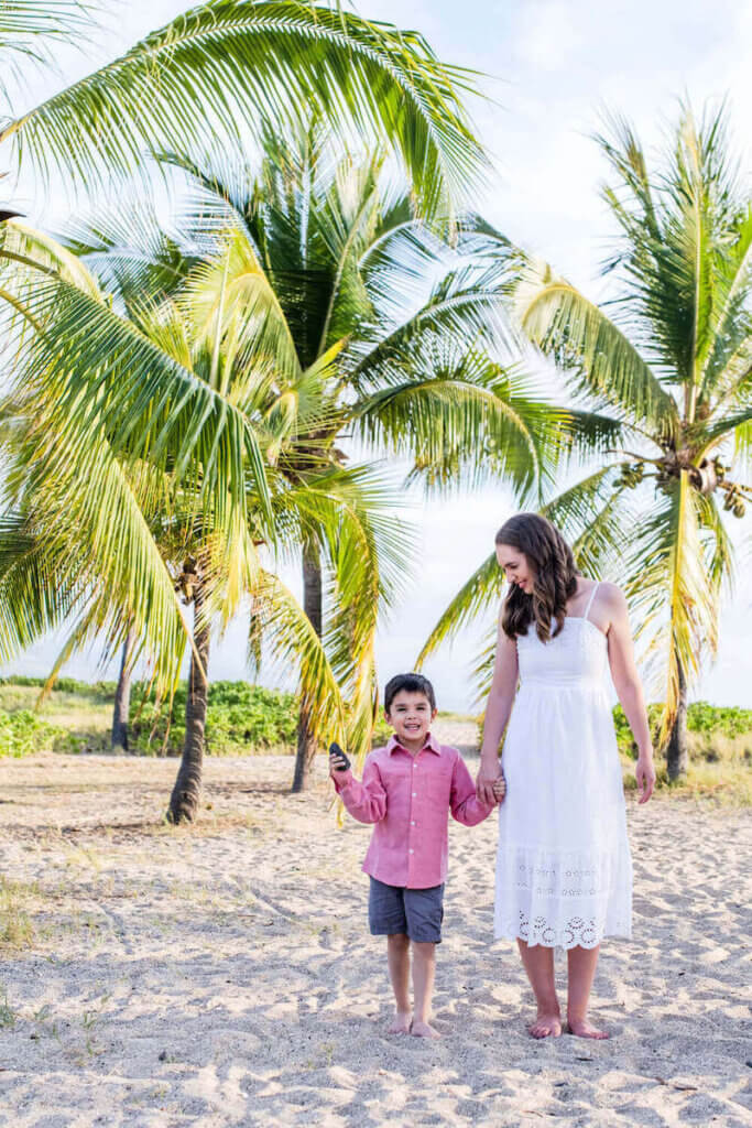 Image of a mom wearing a white dress and a boy wearing a red shirt walking on the sand in Hawaii.