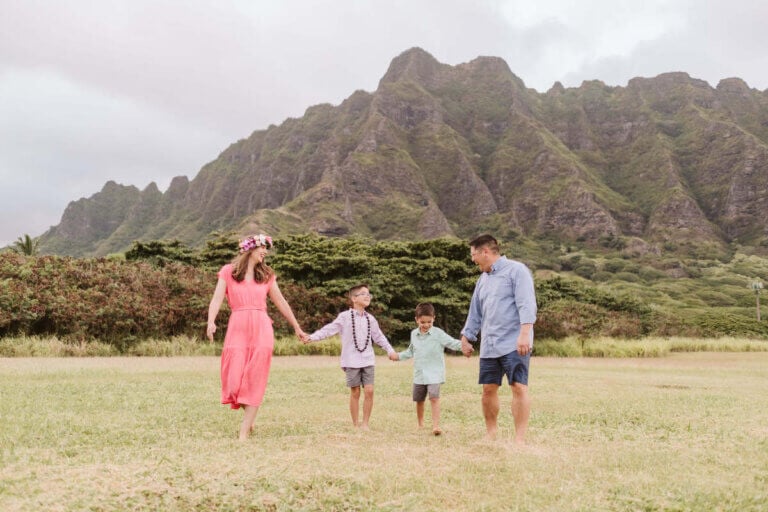 Find out how to book family photos in Hawaii by a mom who's done it on Oahu, Maui, Kauai, and the Big Island! Image of a family of four holding hands with Kualoa Mountains in the background