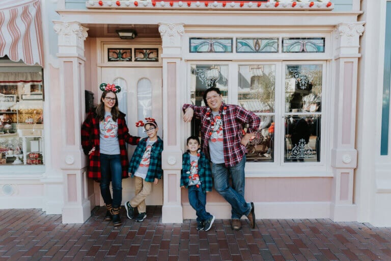 Find out how to book a Disneyland photography session by top family travel blog Marcie in Mommyland. Image of a family wearing Christmas Mickey and plaid in front of a shop on Main Street at Disneyland California.