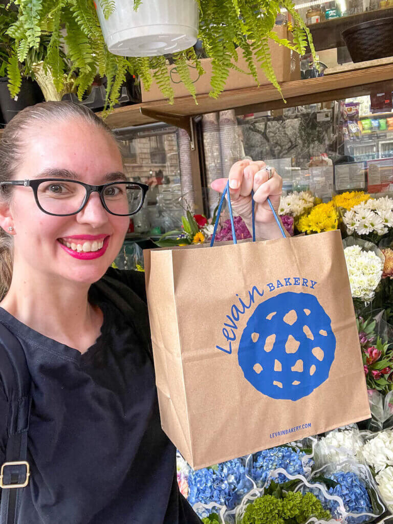 Image of a woman holding a Levain Bakery bag with flowers in the background