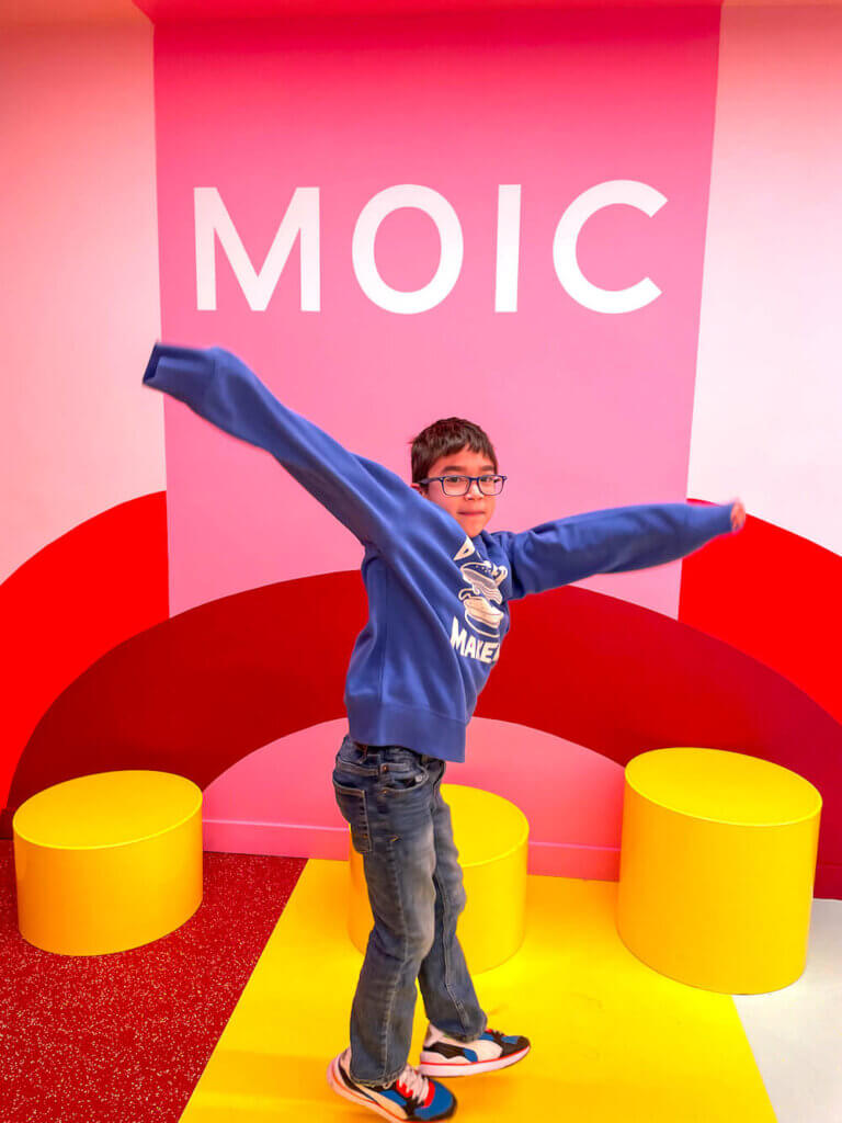 Definitely add the Museum of Ice Cream to your New York in 7 Days Itinerary. Image of a boy standing in front of a colorful wall with MOIC on it