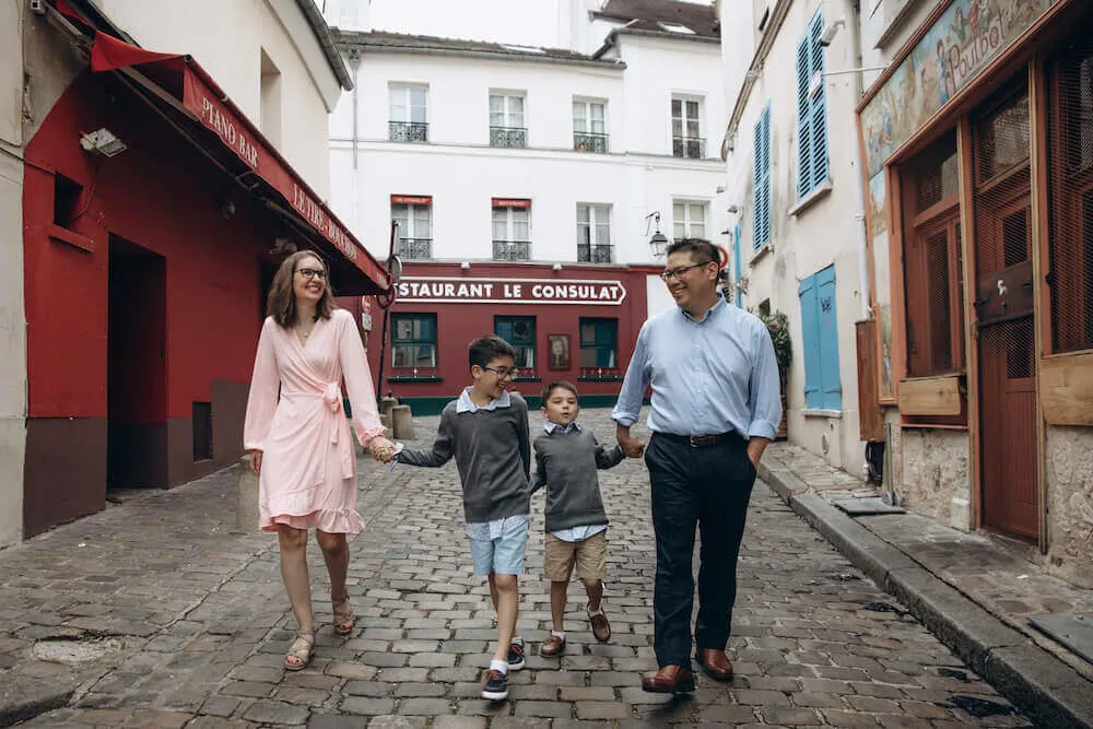 Image of a mom wearing a pink dress, two boys wearing grey sweaters and shorts, and a dad wearing a blue button down shirt and slacks walking down a street in Paris.