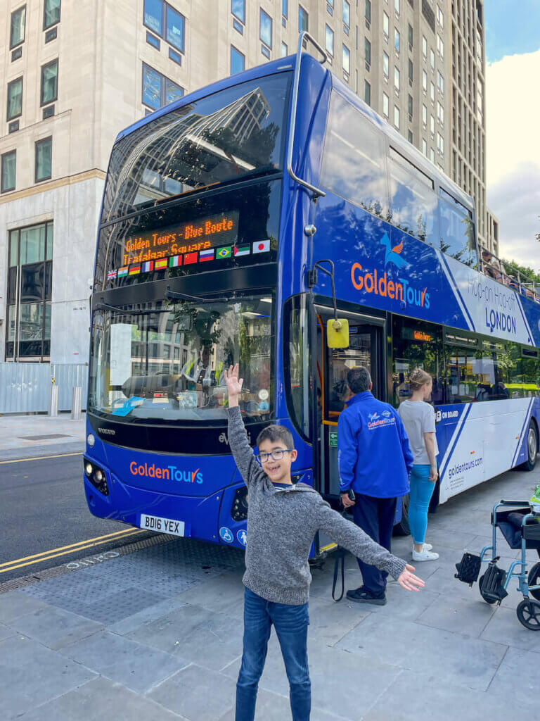 Image of a boy with outstretched arms in front of a blue double decker bus in London
