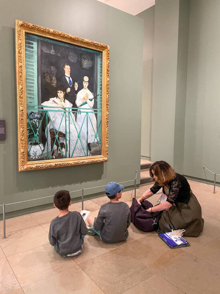 One of the best Paris tours for families is this scavenger hunt at the Musee d'Orsay. Image of a woman helping two kids paint in front of a painting