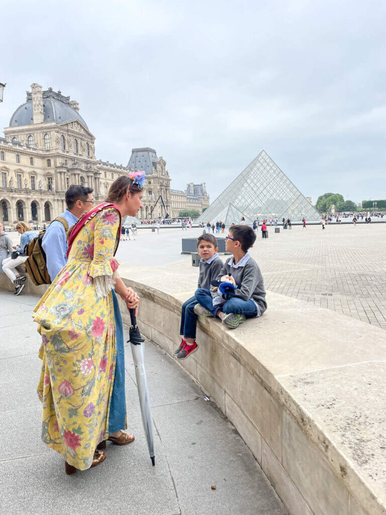 Image of a woman dressed in 1700s costume with two little boys in front of the glass pyramid at the Louvre in Paris