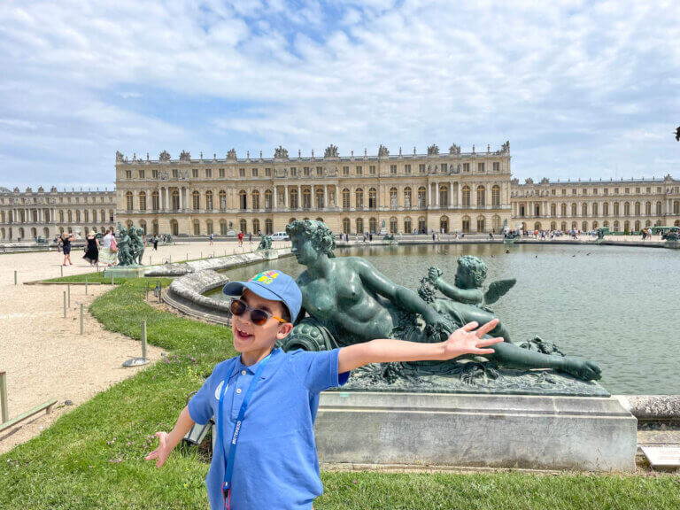 Find out whether or not it's worth visiting Versailles with kids. Image of a boy with outstretched arms in front of a statue at the Palace of Versailles