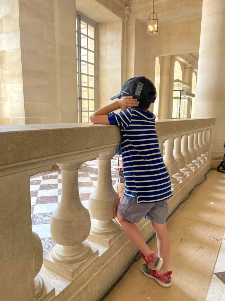 Image of a boy with his hands over his ears leaning up against a railing inside the Palace of Versailles.