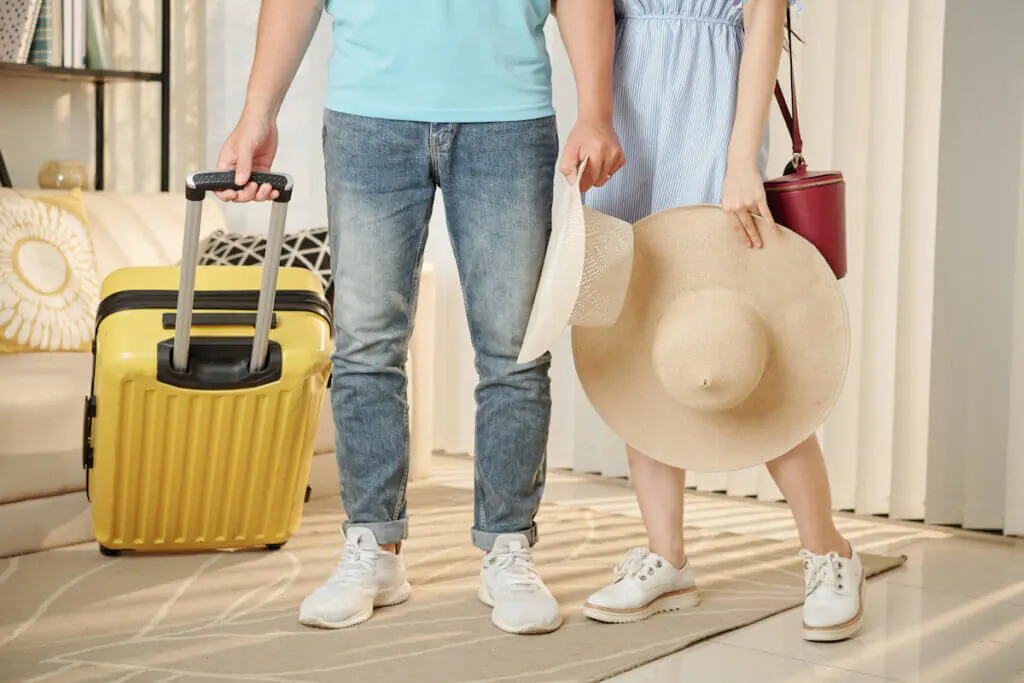 Low section of young couple with straw hats standing in room with suitcase ready for summer vacation