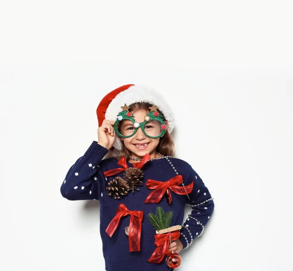 Cute little girl in handmade Christmas sweater and hat with party glasses on white background