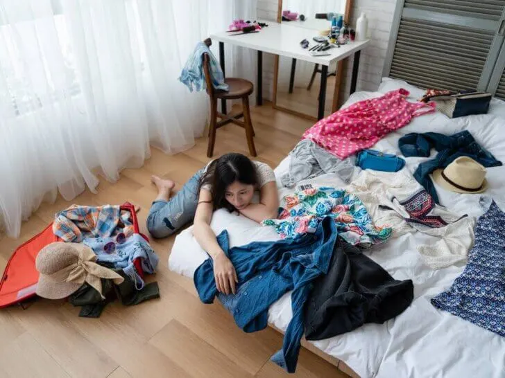 Find out the worse packing mistakes families make, according to top family travel blog Marcie in Mommyland. Image of top view of young asian woman at home packing for luggage in bedroom alone relying thinking daydreaming with head leaning on messy white bed with clothes dresses for vacation. female prepare suitcase