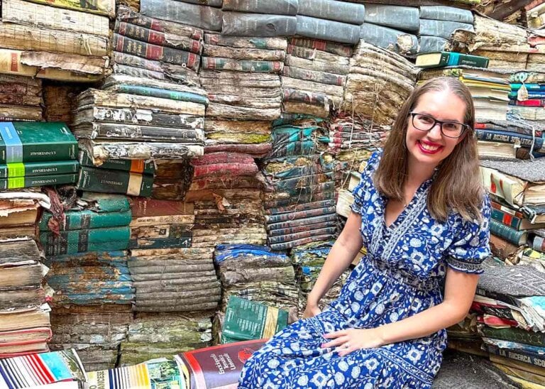 Check out these amazing Venice hidden gems recommended by top family travel blog Marcie in Mommyland. Image of a woman sitting next to a bunch of books at the Cat Library in Venice
