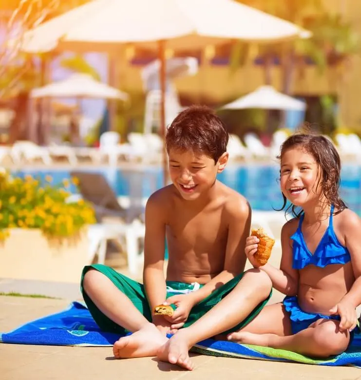 Image of Two happy kids eating croissant near pool, having breakfast on the beach, active summer holidays, brother and sister enjoying sunny day, happiness concept