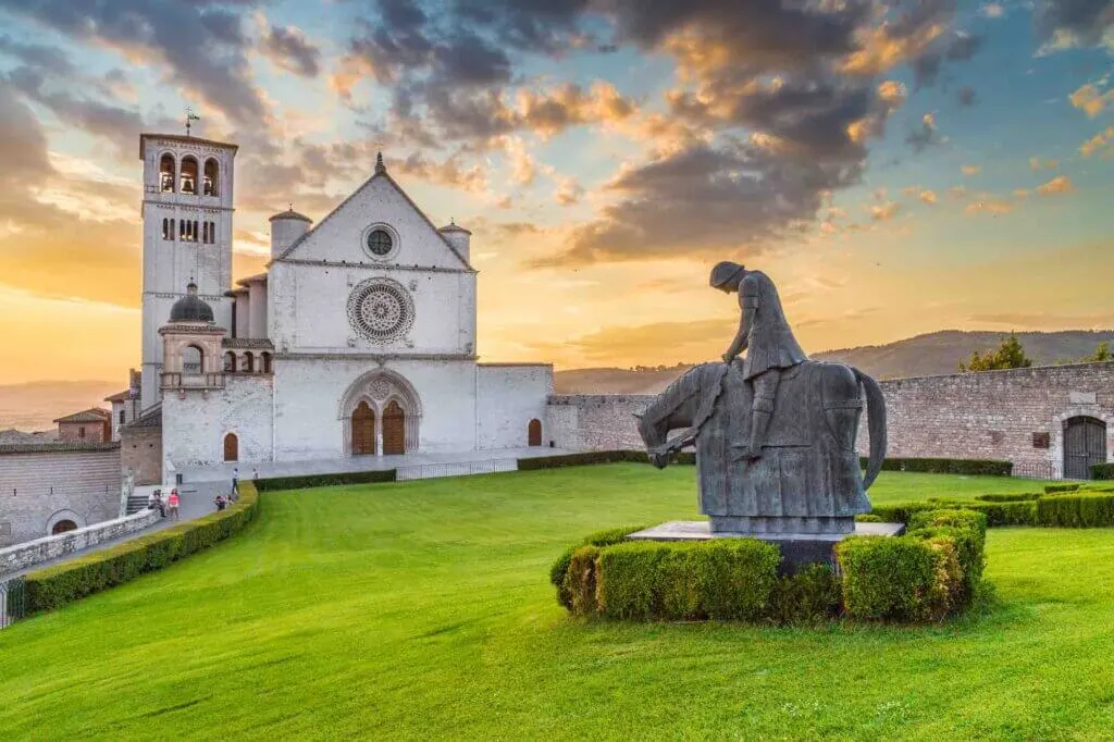 Classic view of famous Basilica of St. Francis of Assisi (Basilica Papale di San Francesco) with statue in beautiful golden evening light with dramatic clouds in the sky at sunset, Assisi, Umbria, Italy