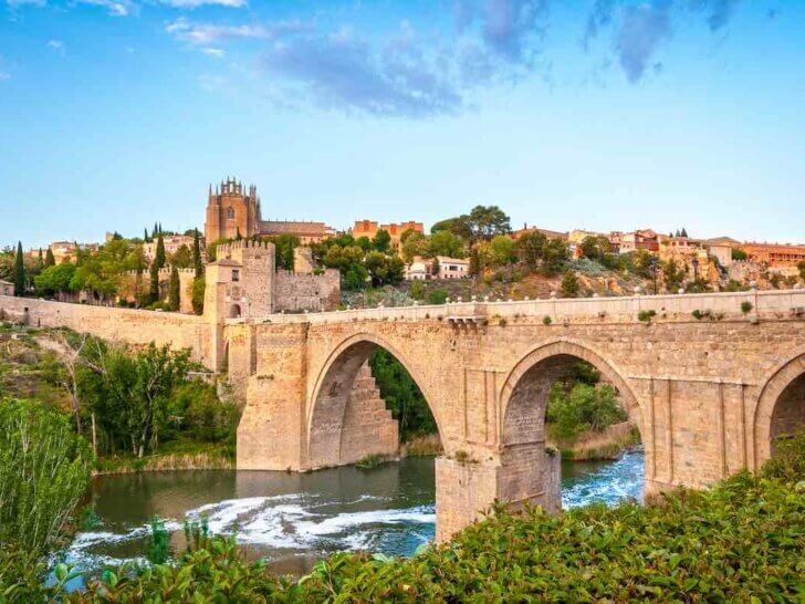 Check out the best day trips from Madrid by train recommended by top family travel blog Marcie in Mommyland. Image of Beautiful landscape of Toledo in Spain. Stone bridge across calm river. Blue sky reflected in crystal clear water. Big fort and country houses in background. Popular tourist place in Europe.