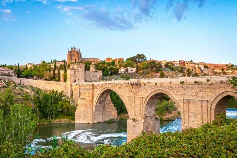 Check out the best day trips from Madrid by train recommended by top family travel blog Marcie in Mommyland. Image of Beautiful landscape of Toledo in Spain. Stone bridge across calm river. Blue sky reflected in crystal clear water. Big fort and country houses in background. Popular tourist place in Europe.
