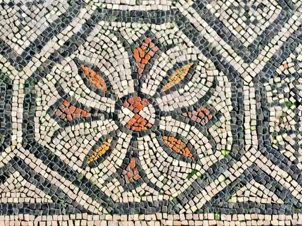 A roman mandala mosaic executed on an early roman floor, showing a syliosed flower in a heaxagon format with vivd reds and oranges