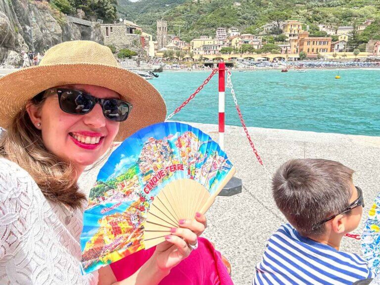 Find out how to do a day trip to Cinque Terre with kids with tips by top family travel blog Marcie in Mommyland. Image of a woman and a boy holding fans while sitting at the pier in Monterosso al Mare