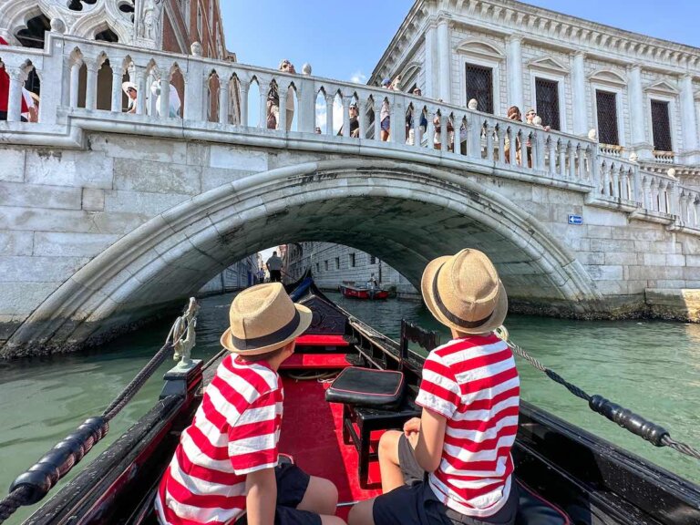 Find out how to spend 1 day in Venice with kids with tips by top family travel blog Marcie in Mommyland. Image of two boys wearing red and white striped shirts in a gondola in Venice Italy