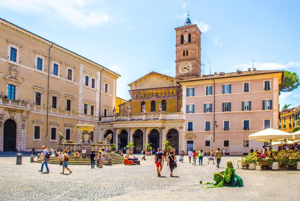 people are strolling through piazza di santa maria situated in front of the basilica with the same name in trastevere district in rome.