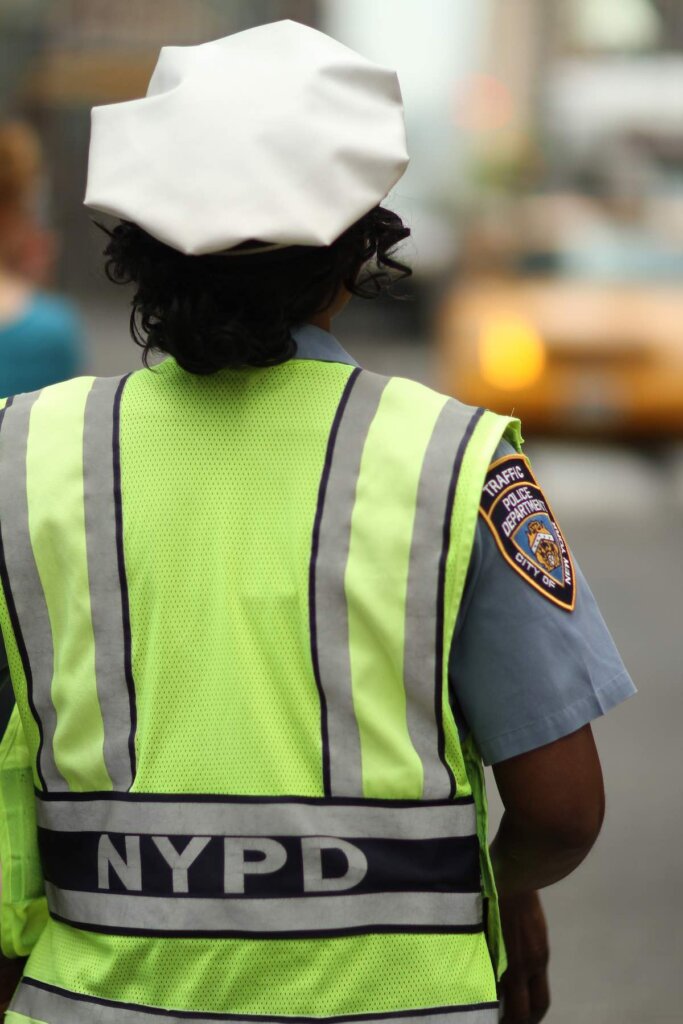An African American policewoman monitors traffic in a city environment, wearing a yellow vest and a white hat, her badge clearly showing on sleeve.