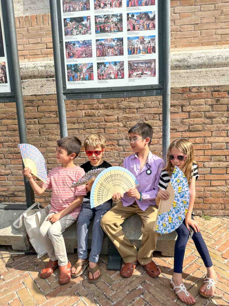 Image of four kids holding fans while sitting beneath a sign at the Vatican