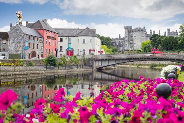 Check out these day trips from Dublin by train recommended by top family travel blog Marcie in Mommyland. Image of beautiful flower lined riverside view of kilkenny castle town and bridge