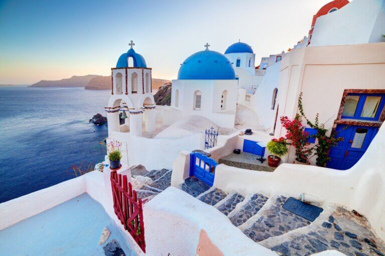 Find out the best Santorini hotels for families recommended by top family travel blog Marcie in Mommyland. Image of Oia town on Santorini island, Greece at sunset. Traditional and famous churches with blue domes over the Caldera, Aegean sea