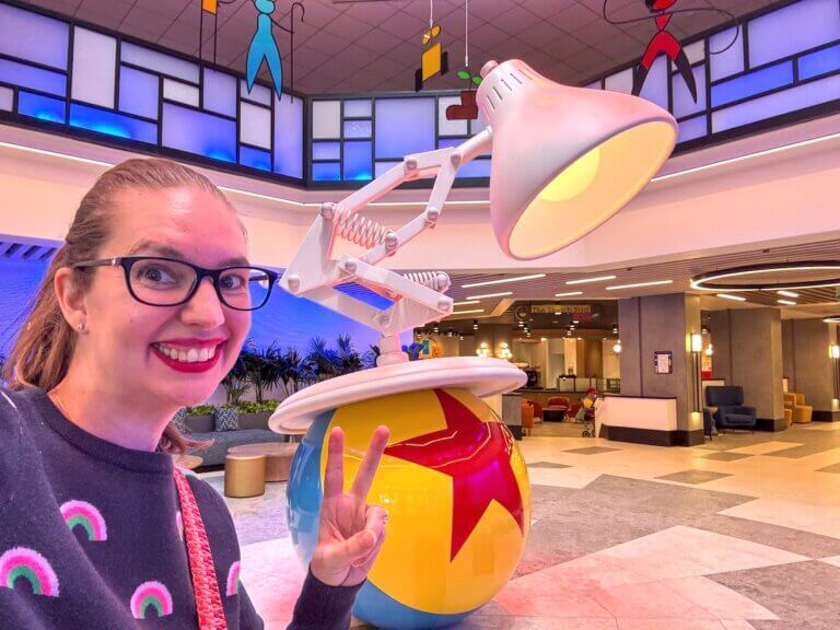 Check out this honest Pixar Place Hotel Review by top family travel blog Marcie in Mommyland. Image of a woman in front of the Pixar lamp in the lobby of the Pixar Place Hotel at Disneyland.